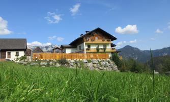 A beautiful house with a balcony, surrounded by a green meadow and mountains in the background. The sky is blue with some white clouds.