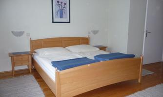 A cozy bedroom with a large double bed and wooden furniture. Pictures hang on the walls, and the floor is covered with a carpet.