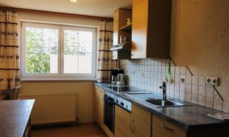 A modern kitchen with wooden cabinets and a large window. The room is well-lit and has a practical arrangement of appliances and work surfaces.