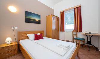 A bright and cozy bedroom with a double bed, a nightstand, and a desk. While a picture on the wall and curtains beautify the room, a window provides natural light.