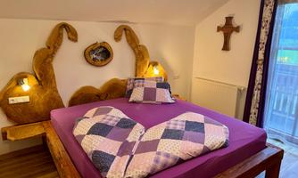 A cozy bedroom with a large bed and wooden details. The ceilings are striped in purple and gray, and the room has a window opening to the balcony.