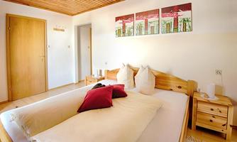 A bright bedroom with a double bed and two decorative pillows. On the wall, three pictures are visible and a wooden door frame is visible.