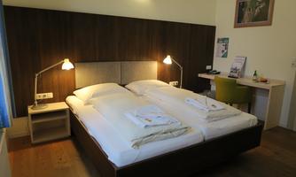 A cozy room with a double bed and two bedside lamps. Next to the bed is a desk with a chair and some information for guests.