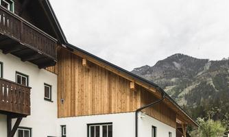 A modern house with a wooden section and large windows. In the background, mountains and a cloudy sky can be seen.
