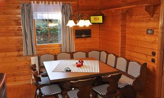 A cozy dining area made of wood with a large table and many chairs. Windows with curtains and soft lighting create a warm atmosphere.