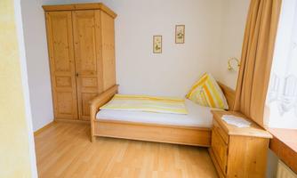 A simple room with a single wooden bed and a light wood chest of drawers. The walls are painted in soft colors, and there is a window with curtains.