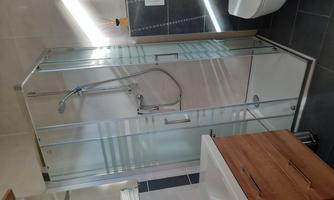A modern bathroom with a glass shower and a wooden sink. The walls are light and the tiles are dark.