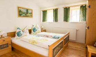 A cozy bedroom with a double bed and light wood furniture. The windows are decorated with green curtains and a painting hangs on the wall.