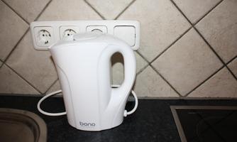 A white kettle stands on a countertop in a kitchen. The walls are adorned with coordinating tiles.