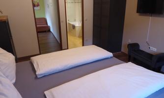 A simple and modern room with two beds and a TV. Through the door, you can see another living area and a bathroom.