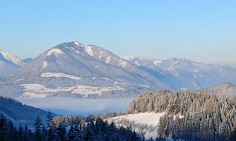 A tranquil winter landscape with snow-covered mountains and forests. The sky is clear and blue, while fog hovers in the valleys.