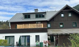 A modern house with two stories and a wooden porch. The garden is green and well-maintained, surrounded by mountains.