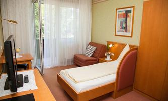 A simple single room with a bed, a desk, and a comfortable armchair. The room has large windows that let in plenty of daylight.