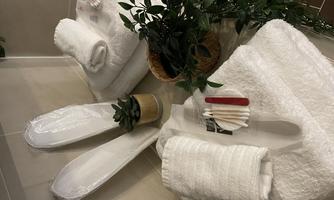 White towels and bath shoes are on a shelf. Next to them is a pot with a plant and some utensils.