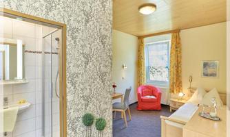 A bright room with a comfortable red armchair and a table. On the left side, there is a bathroom with a shower.