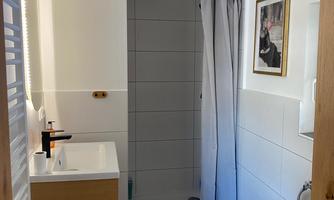 A modern bathroom with a shower, a sink, and a toilet area. The walls are clean and bright, and there is a large window that lets in natural light.