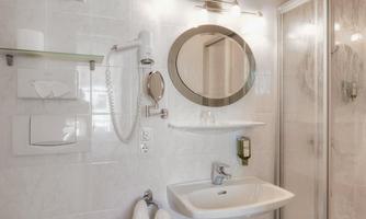A modern bathroom with bright tiles, a sink, and a round mirror. The shower cabin is also visible, along with towels and basic toiletries.