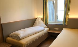 A simple bedroom with two single beds and a window. The walls are light and the curtains are colorful.