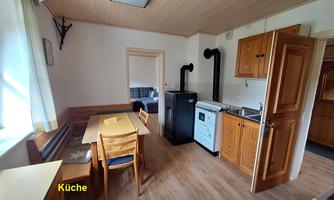 A cozy kitchen with wooden cabinets, a table, and chairs. In the background, other rooms are visible.