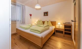 A cozy bedroom with a double bed and green cushions. The room is bright and friendly, featuring wooden floors and warm lighting.