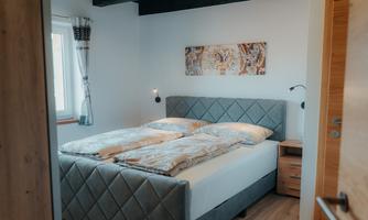 A cozy bedroom with a modern double bed and bright walls. Next to the bed is a nightstand, and a piece of artwork adorns the wall.