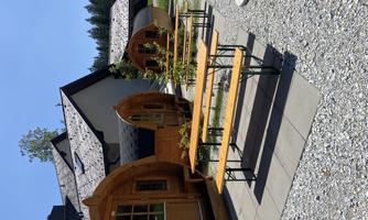 Two cozy wooden cabins stand on a paved square. In the foreground, there are benches and tables outdoors under a clear blue sky.