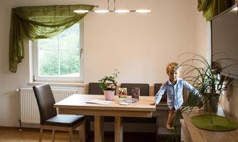 A modern dining room with a table and chairs. Next to the table stands a doll and there are plants on the table.