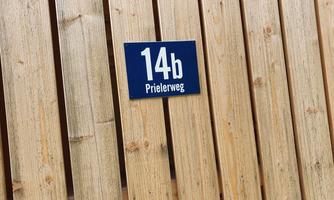 A wooden fence with a blue house number sign displaying the number 14b and the street name "Prielerweg". The background consists of vertical wooden slats.