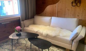 A cozy room with a bright sofa on which a white blanket lies. Next to the sofa is a small table with a flower in a vase.