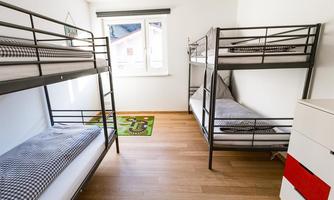 A simple bedroom with two bunk beds and a large window. The floor is made of wood and there is a carpet on the floor.