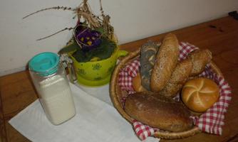 A basket with various rolls and bread is placed on a white cloth. Next to the basket is a glass of milk and a small green bowl with a flower.