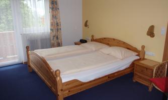 A cozy bedroom with a large wooden double bed. The room has carpet flooring and large windows with curtains.