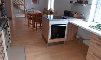 A modern kitchen with wooden furniture and a clean, tidy design. In the background, there is a dining area with a table and chairs.