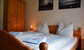 A cozy bedroom with a wooden bed and two pillows. Pictures hang on the wall and a wardrobe stands next to the bed.