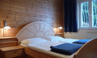 A cozy bedroom with wooden paneling and a large double bed. Next to the bed are nightstands with lamps and a window with curtains.