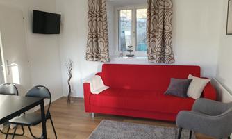 A cozy living room with a red sofa and a gray rug. There is a table with chairs and a television on the wall.