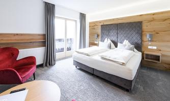 A modern room with a large double bed and a cozy red armchair. There are large windows that let in plenty of daylight.