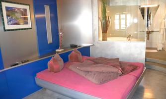 A modern bedroom with a large, comfortable bed in pink and gray colors. The walls are painted blue, and there are decorative elements like a painting and plants.