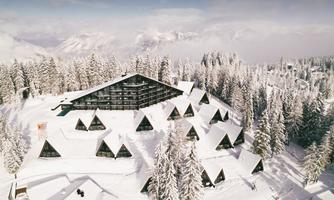 A picturesque mountain landscape with snow-covered trees and modern chalets. In the background, there is a large, modern building.
