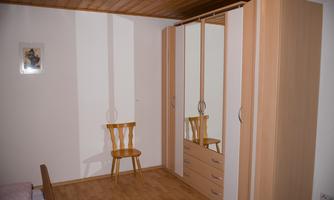 A simple room with a wooden chair and a large wardrobe. The walls are light and the floor is made of parquet.