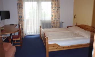 A simple room with a double bed, a table, and a television. The room has large windows with curtains and is bright and friendly.