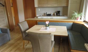 A cozy dining area with a wooden table and upholstered chairs. In the background, there is a modern kitchenette and a houseplant.