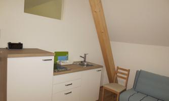 A cozy room with a small kitchenette and a blue sofa. The walls are plain and the ceiling has visible wooden beams.