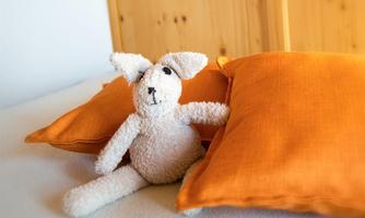A cute plush toy with big ears sits between two orange pillows. The background is bright and cheerful.