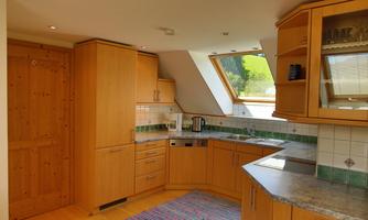A modern kitchen with wooden cabinets and a large window area. The room is bright and inviting with a colorful rug.