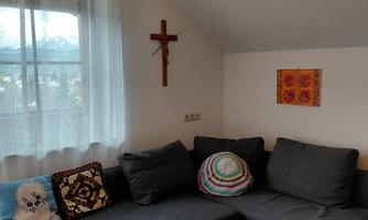 A cozy living area with a gray sofa and a wooden table. On the wall hang curtains and decorations, including a cross and a colorful picture.