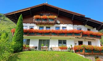 A charming house with a wooden gable, surrounded by green meadows. The balconies are decorated with colorful flowers and the sky is clear and blue.