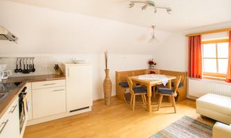 A bright, cozy kitchen with a dining table and chairs. The room has large windows with orange curtains and a warm wooden floor.