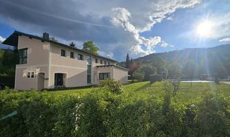 A modern house surrounded by green garden areas. In the background, hills and a sunny sky can be seen.