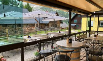 A cozy outdoor area of a restaurant with a table and chairs. In the background, wooden buildings and a garden can be seen.